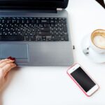 A woman working at a home office desk, with a laptop, cell phone, and coffee.