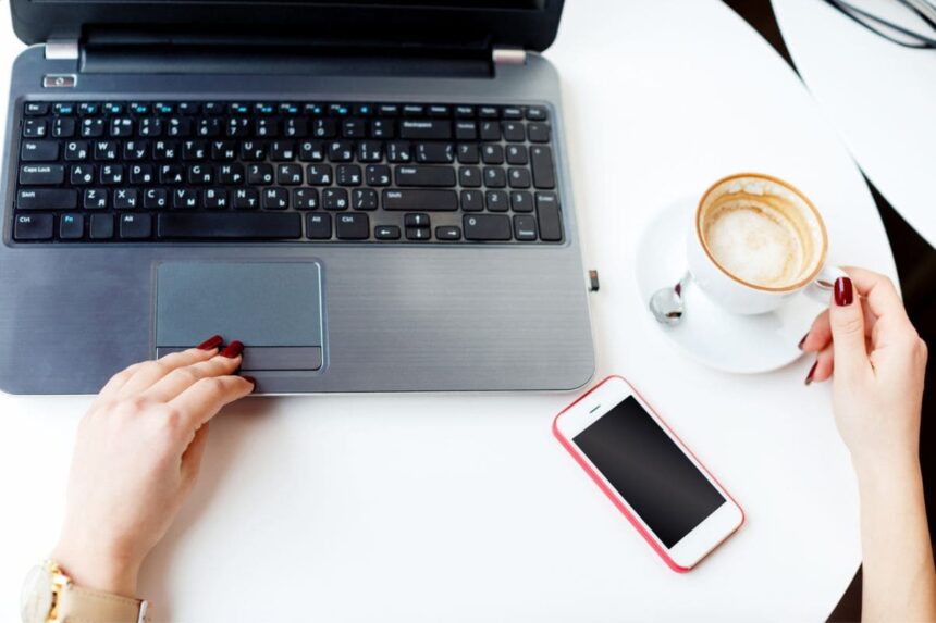 A woman working at a home office desk, with a laptop, cell phone, and coffee.