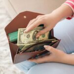 A woman, holding a wallet and putting a $5 bill into it.