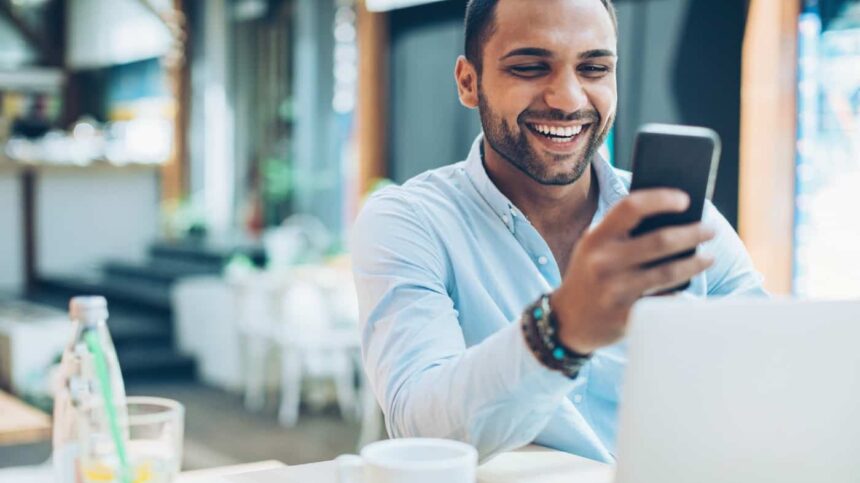 Smiling young man sitting in cafe and checking messages, with his laptop in front of him.