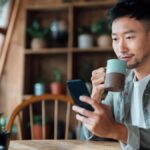 Young Asian man drinking coffee at home and looking at his phone