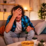 Frustrated young white male looking disconsolate while sat on his sofa holding a beer