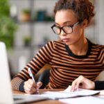 A woman working from home on a business plan, and writing in a notebook.