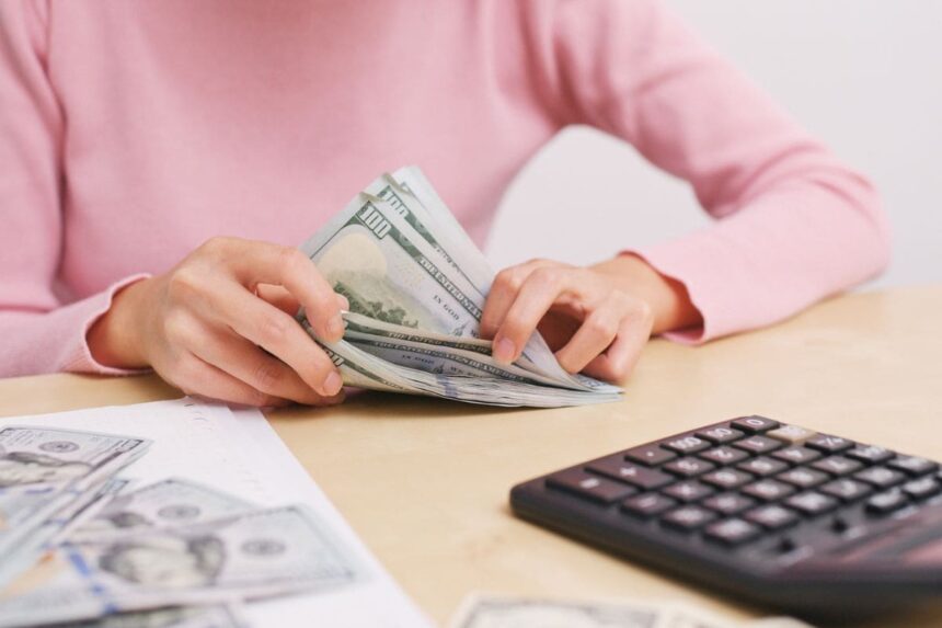 A woman sitting at a desk in her home office, counting cash for her bills and using a calculator.