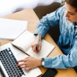 Overhead view of a woman sitting at a home office desk, using a laptop, and writing in a notebook.