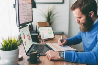 Bearded man writing on notepad in front of computer