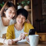 Affectionate Asian senior mother and daughter using smartphone together at home, smiling joyfully