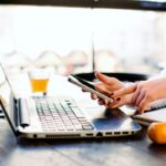 A woman holding a smart phone while working on a laptop at home.