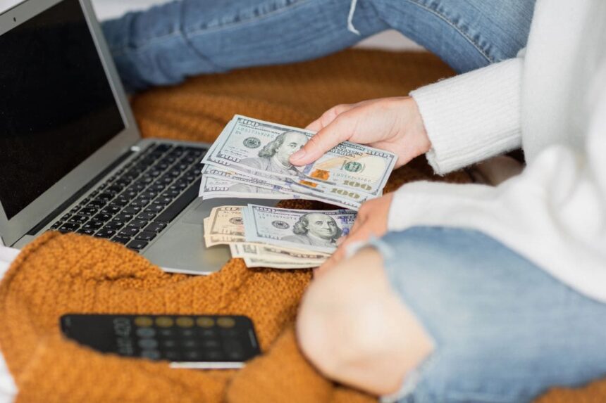 A woman sitting on her living room floor, using a laptop, and counting cash.