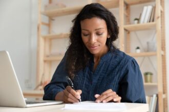 A woman working remotely from her home office, using a laptop and writing in a notebook.