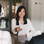 A woman sitting down in an office across from two people for an interview. She's handing them her resume.