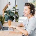 A woman sitting at a home office desk and using a laptop and a headset to do remote dispatcher work.