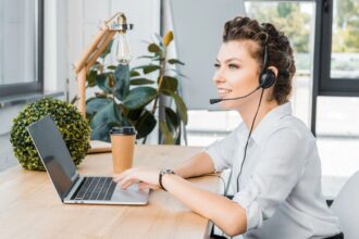 A woman sitting at a home office desk and using a laptop and a headset to do remote dispatcher work.