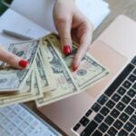 A woman sitting at her home office desk, counting cash while working on a budget.