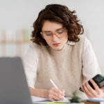 A woman at home sitting at her home office desk, using a laptop and cell phone.