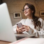 A woman working from a home office, sitting at a desk, holding a phone, and working on a laptop.