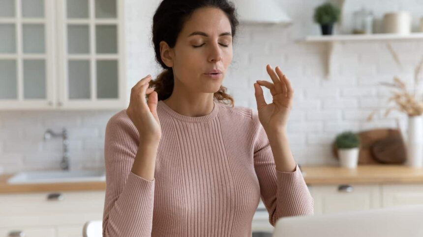 Mindful young woman breathing out with closed eyes, calming down in stressful situation, working on computer in modern kitchen.