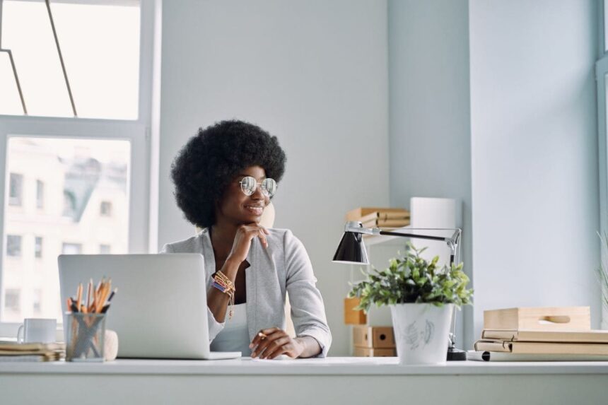 A writer, sitting at her home office desk with a laptop and plant, looking out of the window.