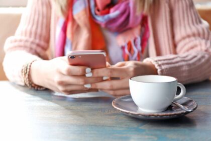 A woman sitting at a table at a cafe, drinking coffee and using her smartphone.