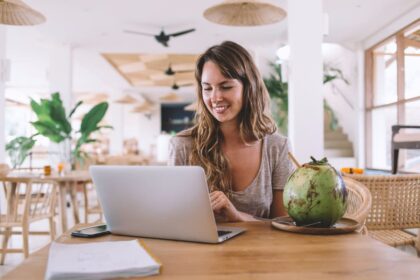 A digital nomad, working remotely on her laptop from a cafe in a tropical location, drinking from a coconut.